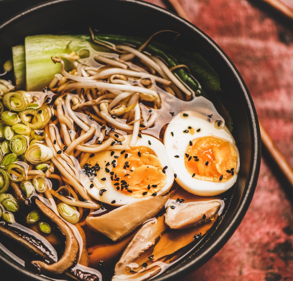 Soupe traditionnelle de ramen japonaise avec viande de poulet et champignons shiitake dans des bols noirs et des baguettes en bambou sur le fond de table en bois rouge foncé, vue de dessus.