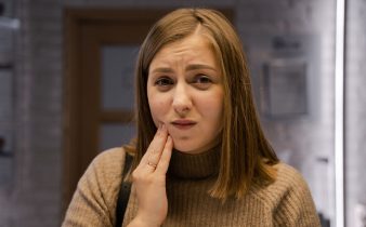 Femme caucasienne exprimant un mal de dents, portant un pull marron à l'intérieur.