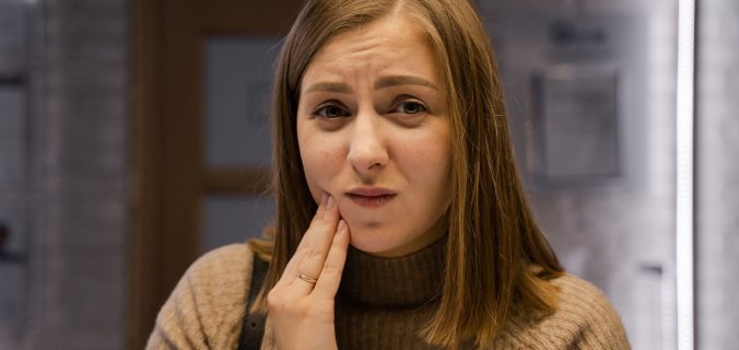Femme caucasienne exprimant un mal de dents, portant un pull marron à l'intérieur.